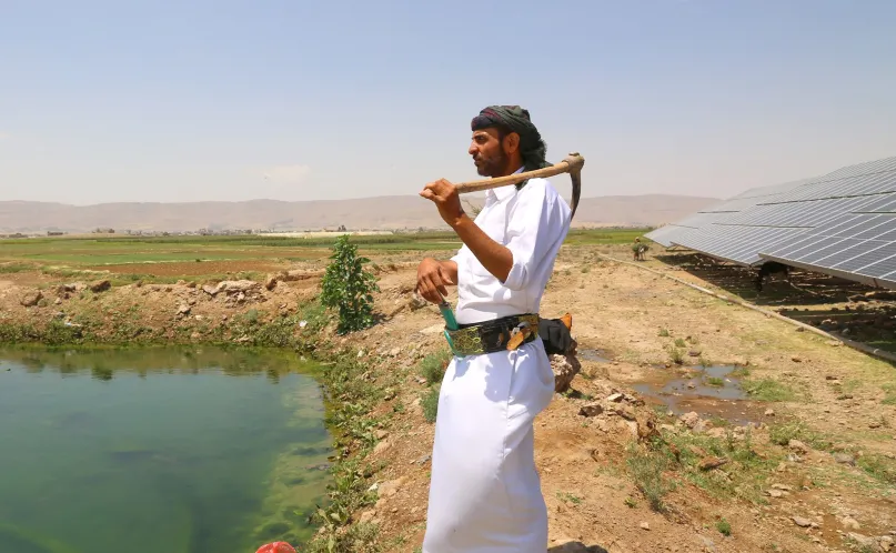 Man in white robe holding pickax over his shoulder, looking out at fields, with solar panels behind him.