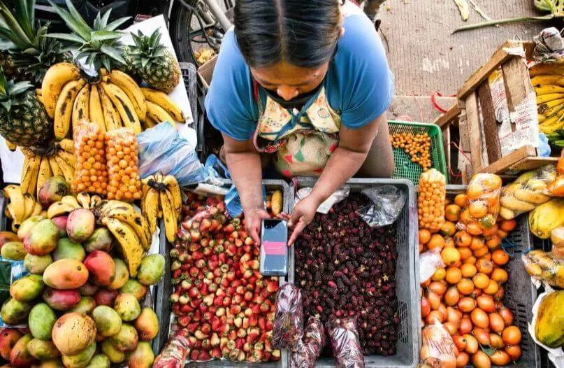 Mujer mirando un celular sobre las frutas y verduras de su puesto en Colombia.