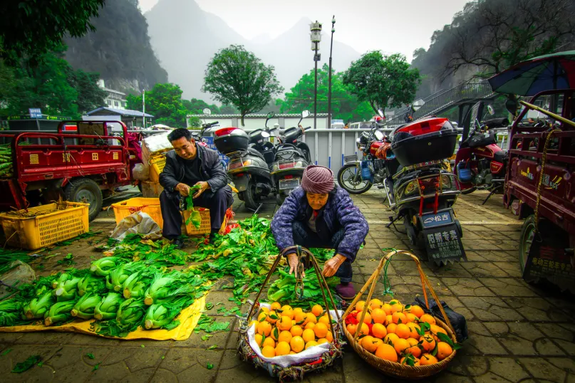 Farmers in China selling vegetables and oranges with motorcycles, green trees and misty mountains behind them.