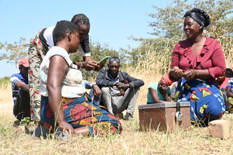 Group of people sitting on a dry field with a cash box looking at a phone. 