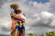 African woman holding baby in field with clouds behind her.