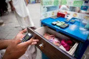 Hand holding a cell phone in front of an open drawer with cash and calculators on the desk.